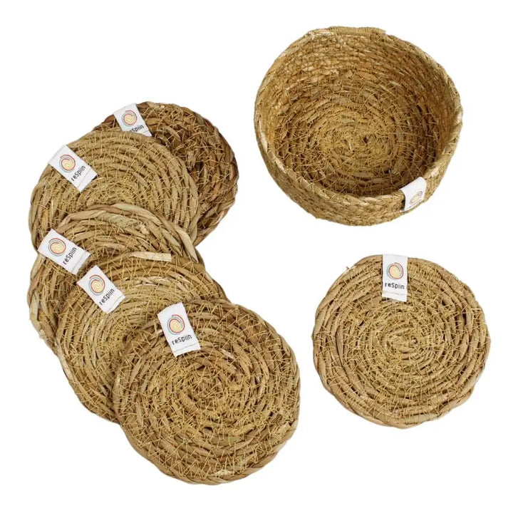 Set of round woven natural fibre baskets and placemats in a light tan straw colour, with some pieces stacked and one basket shown open, on a plain white background. Small white labels with a circular logo are attached to several pieces.
