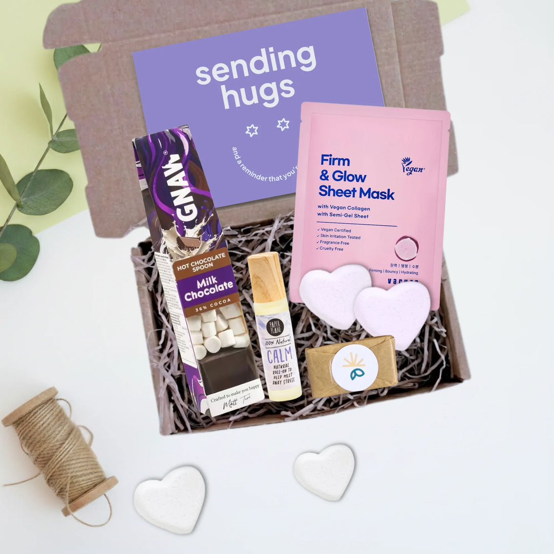 A curated gift box filled with assorted self-care and chocolate treats, photographed from above on a white surface with decorative eucalyptus leaves, twine, and white heart shapes. The box includes a purple card reading &