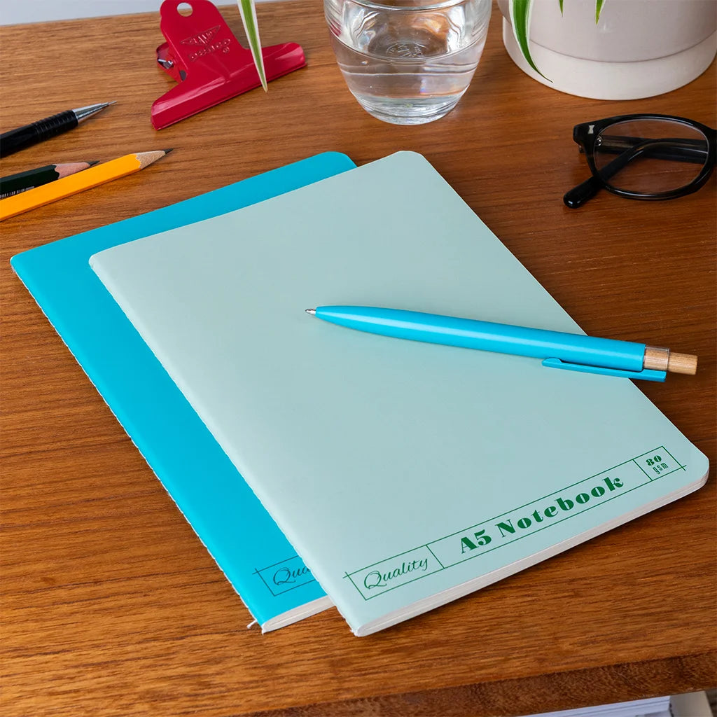 Light blue A5 notebook with a blue pen on a wooden desk with stationery items.