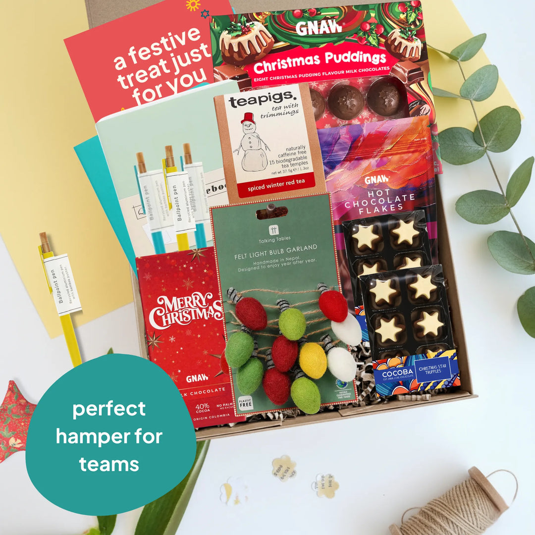 A Christmas-themed team hamper gift box filled with treats and stationery, including GNAW Christmas Puddings, chocolate bars, a light bulb garland, and a Merry Christmas card, arranged on a white surface with decorative leaves and a teal badge that reads &
