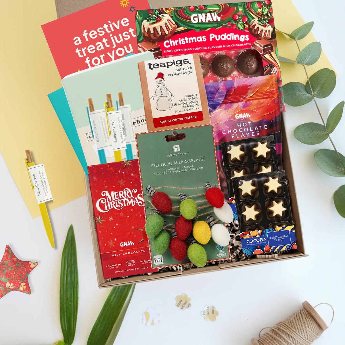 A curated Christmas gift box filled with treats and stationery, photographed from above on a white surface with decorative leaves and paper. Visible items include a red 'Merry Christmas' chocolate bar, a teal 'a festive treat just for you' card, GNAW Christmas Puddings chocolate box, a Teapigs snowman tea bag box, a felt light bulb garland, and chocolate stars.