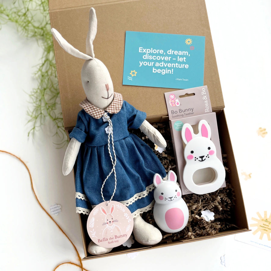 Box containing a plush rabbit toy, baby rattle, white and pink bunny teether and card with text 'explore, dream, discover - let your adventure begin!' on a white background