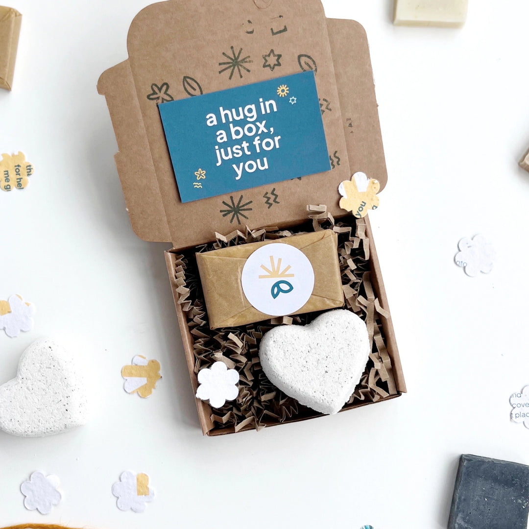 Hug in a box gift including heart-shaped bath bomb and mini soap bar in a printed kraft box with a card inside which reads 'a hug in a box just for you'. The gift box is on on a white background with seed paper flower scattered around.