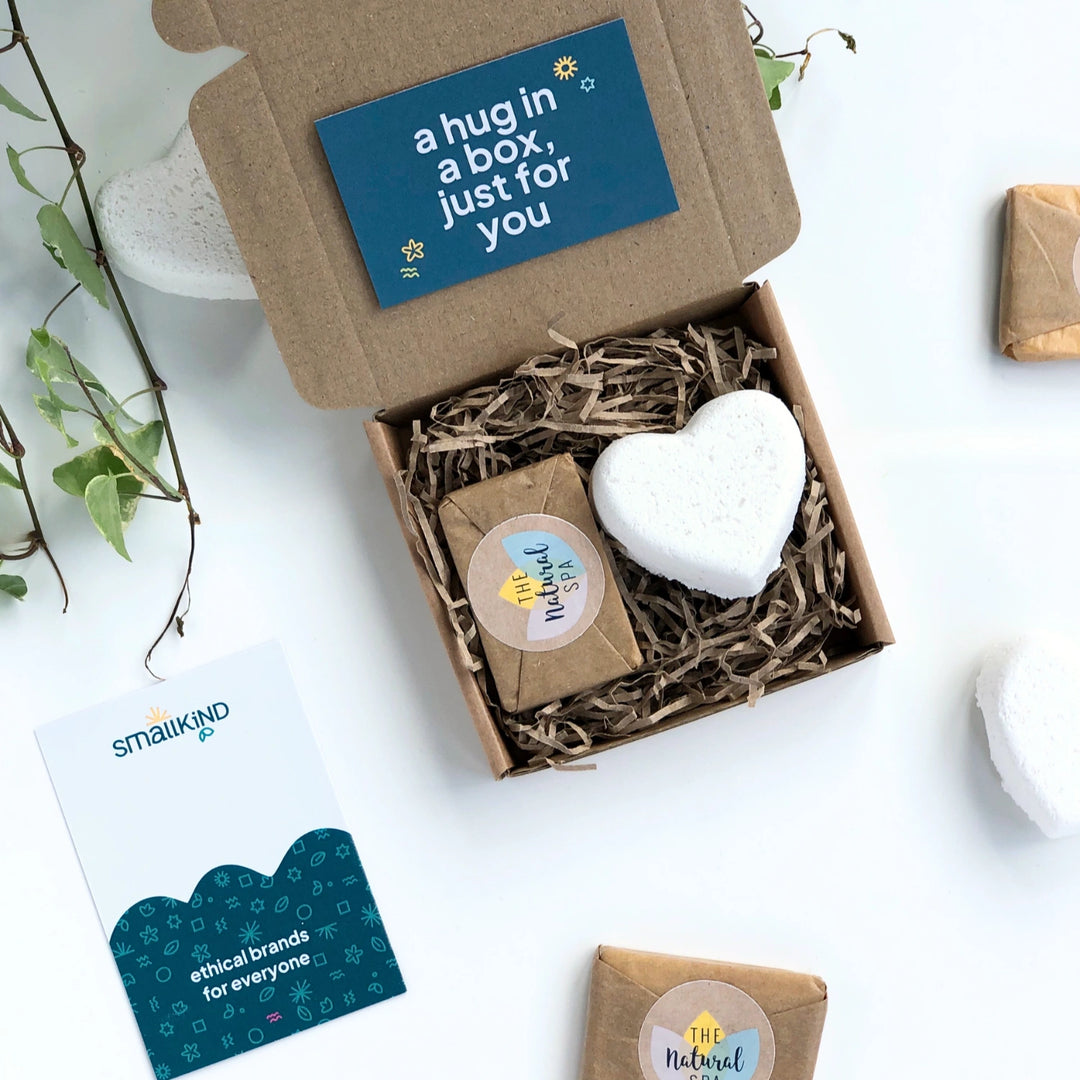 a white heart shaped bath bomb and mini soap wrapped in kraft paper inside a kraft gift box filled with shredded paper. There is a dark green gift card with the words 'a hug in a box just for you' in bold white text