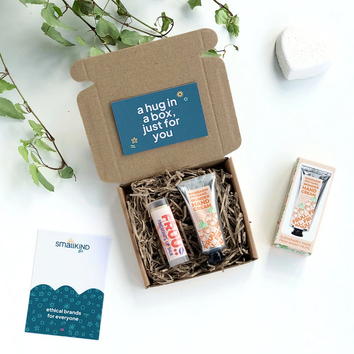 a mandarin orchard hand cream and pomegranate lip balm, both made by Fruu cosmetics, inside a krfat gift box filled with shredded paper. The gift card is dark green and has a message 'a hug in a box just for you' in bold white text. 