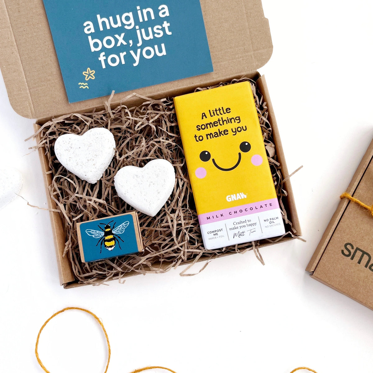Something to Make You Smile Gift Box by Smallkind including two heart shaped bath bombs., a box of seedballs and a milk chocolate bar with a 'a little something to make you smile message'. The gifts are packaged in a hand stamped kraft gift box with shredded paper.