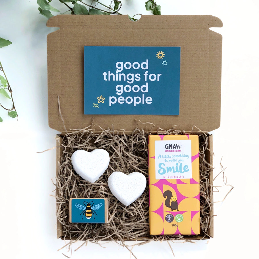 kraft gift box filled with shredded paper containing a chocolate bar with the words 'a little something to make you smile' , two heart shaped bath bombs and a box of wildflower seedballs