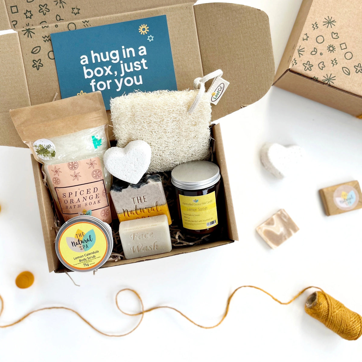a selection of pamper products with a yellow theme including: spiced orange bath soak, a natural bath bomb heart, spiced orange soap bar, face wash bar, lemon sorbet candle in a glass jar. The products are inside a hand stamped gift box with a card which reads 'a hug in a box, just for you'
