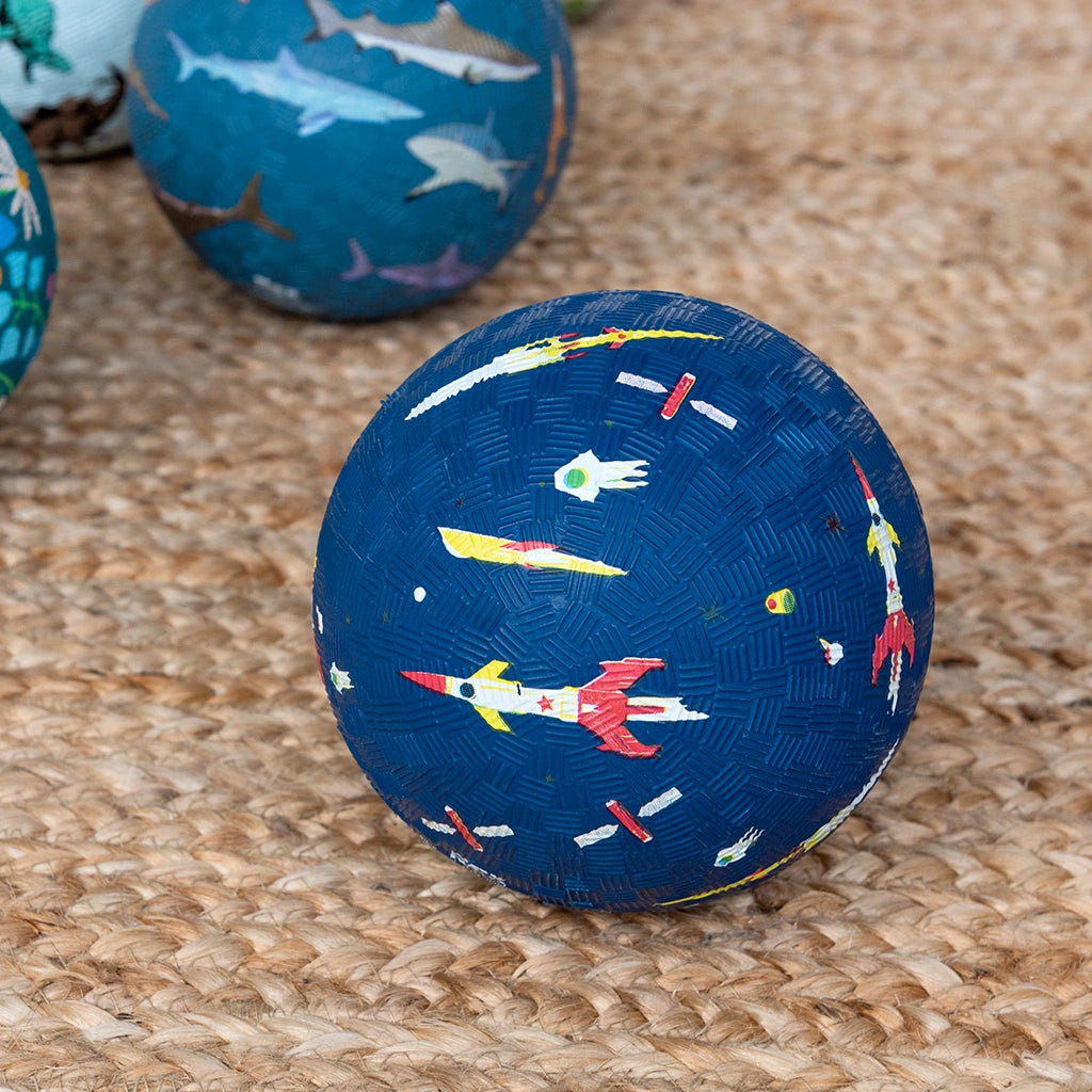 Dark blue rubber play ball with space-themed design featuring rockets and astronauts on a n indoor rug