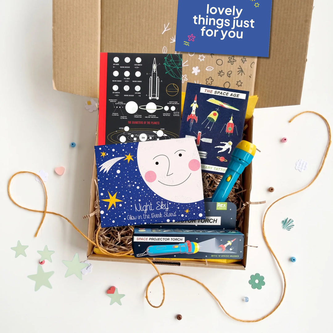 An open cardboard gift box filled with space-themed items, including a space notebook, space rockets temporary tattoos,  glow in the dark stars and a projector torch, arranged on a white surface with decorative star and flower cutouts.