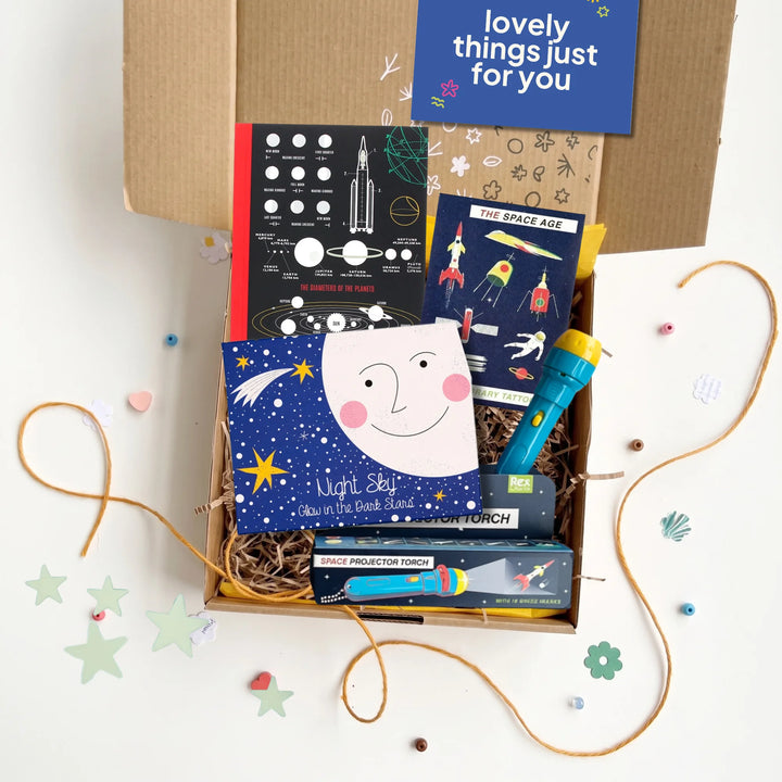 An open cardboard gift box filled with space-themed items, including a space notebook, space rockets temporary tattoos,  glow in the dark stars and a projector torch, arranged on a white surface with decorative star and flower cutouts.