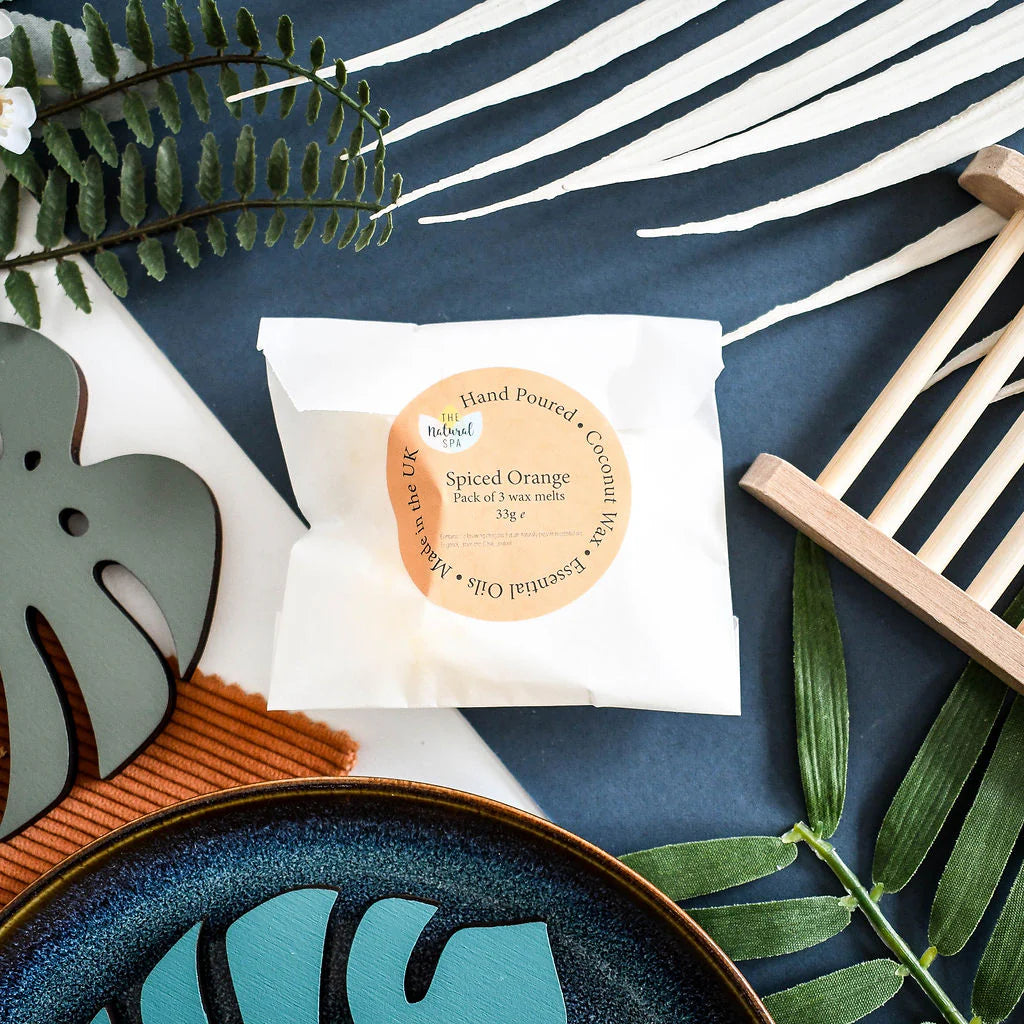 Hand-poured wax melts in a white compostable bag with a decorative label on a blue surface with green leaves and a wooden soap dish.