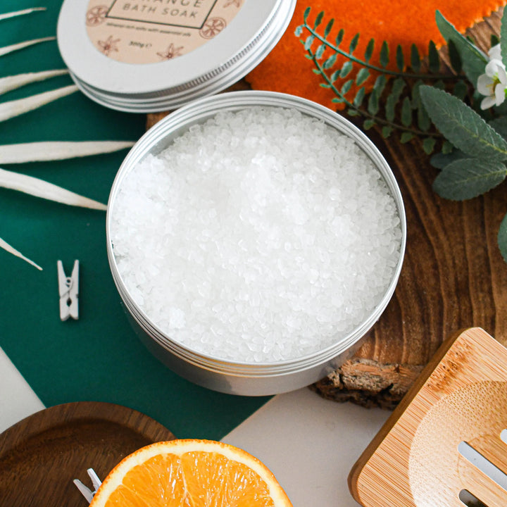 An open round metal tin filled with white bath salts or bath bomb granules, styled on a wooden surface with green leaves, an orange slice, a small clothespin, and stacked tins in the background.