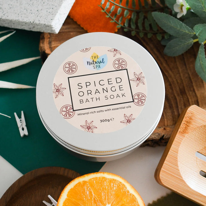 A round tin of The Natural Spa Spiced Orange Bath Soak, styled on a wooden board with an orange slice, green leaves, and a teal background.