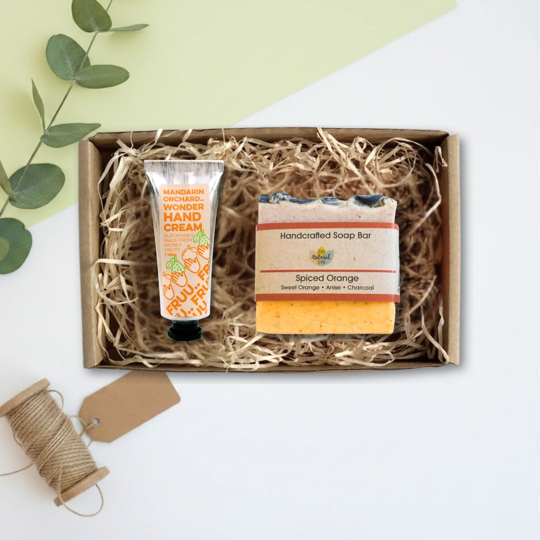 A gift set containing a tube of hand cream and a soap bar, presented in a brown cardboard box filled with shredded paper, on a white background with decorative eucalyptus leaves and a spool of twine.