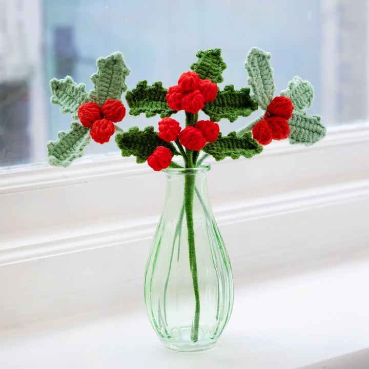 Crocheted holly branches with red berries in a clear glass vase on a windowsill.