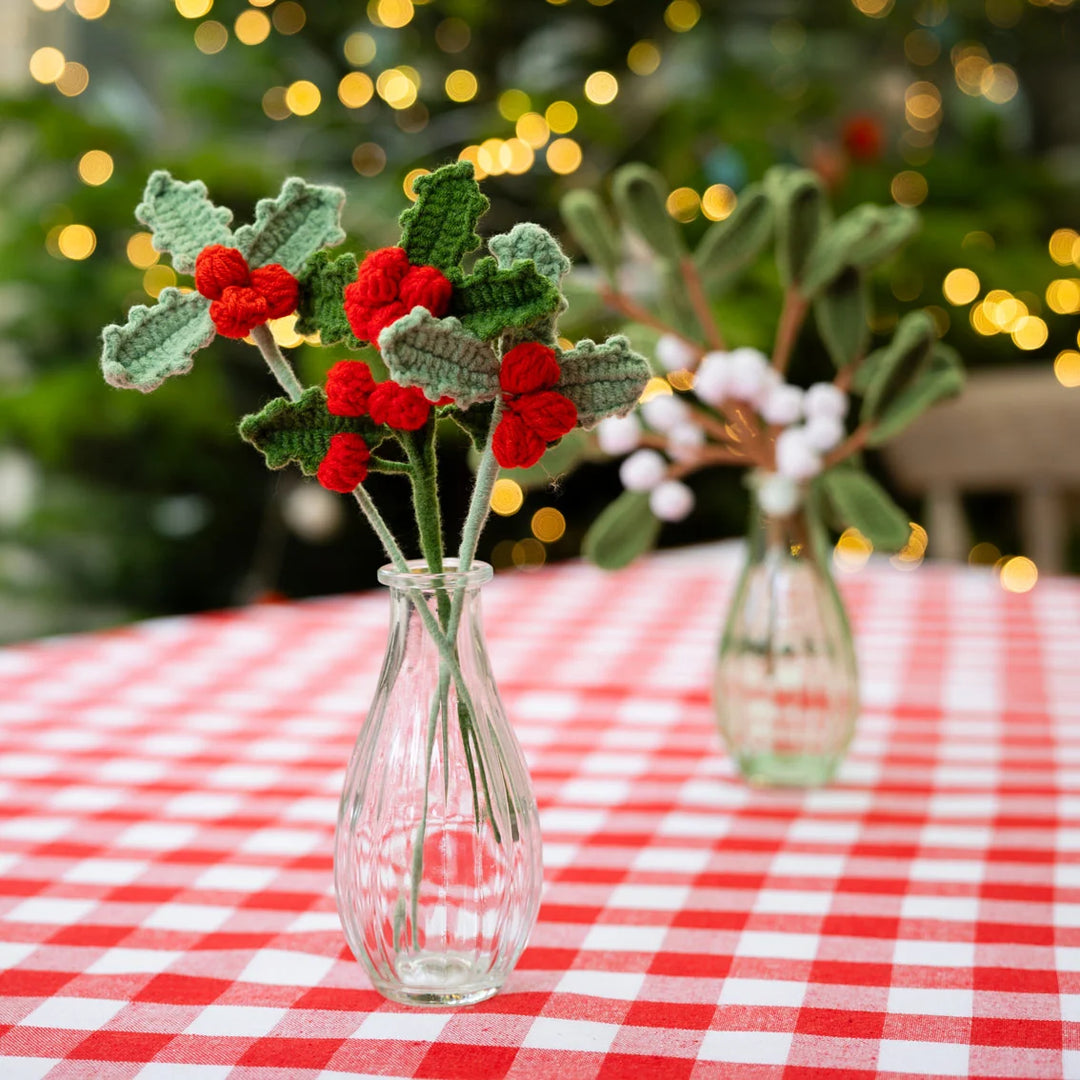 set of 3 crochet holly sprigs in a clear glass bud vase with green foliage in the background.