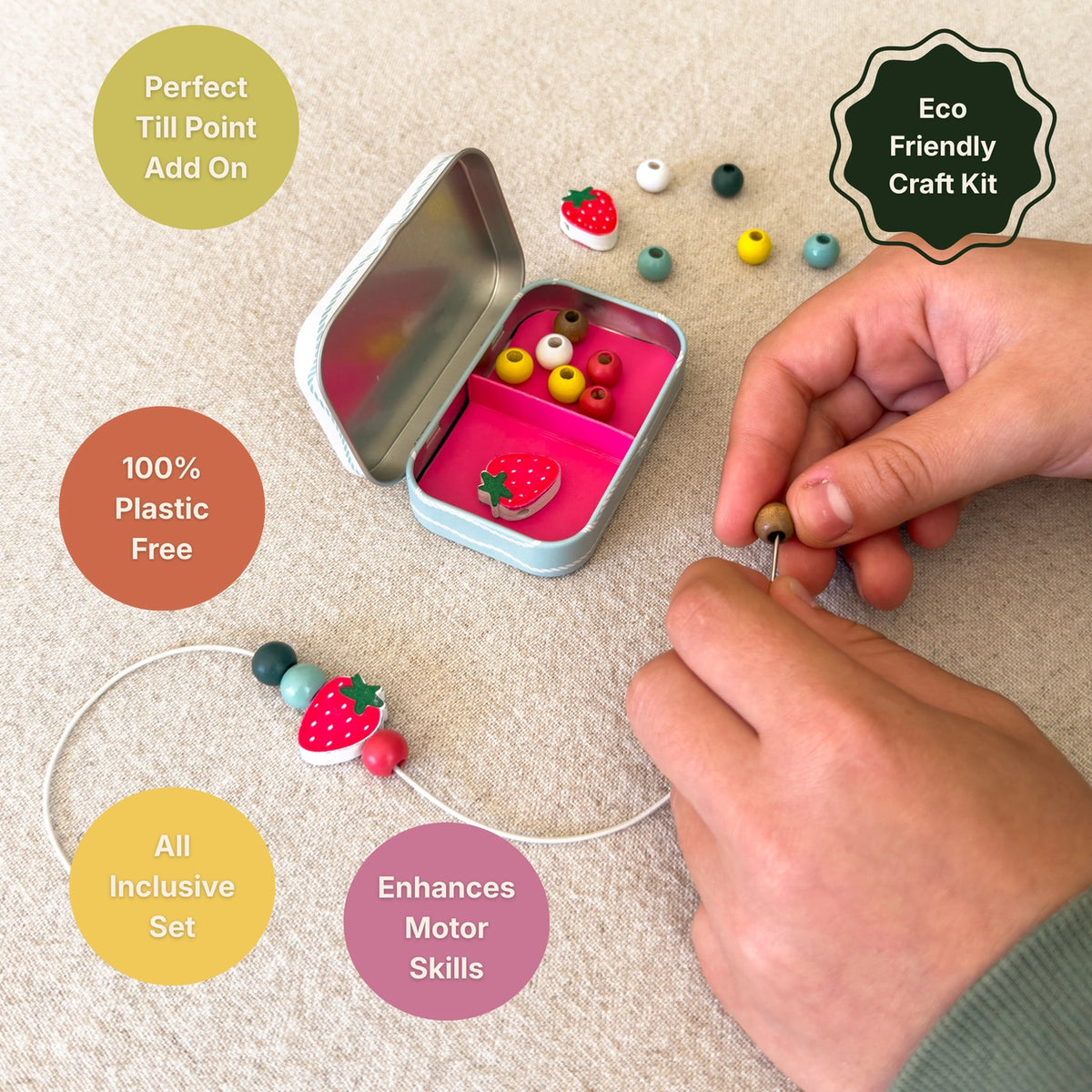 A hands-on craft kit scene showing a person threading beads onto a string. The kit includes a small tin of colorful wooden beads, loose beads, and a completed bead strand with a strawberry bead.