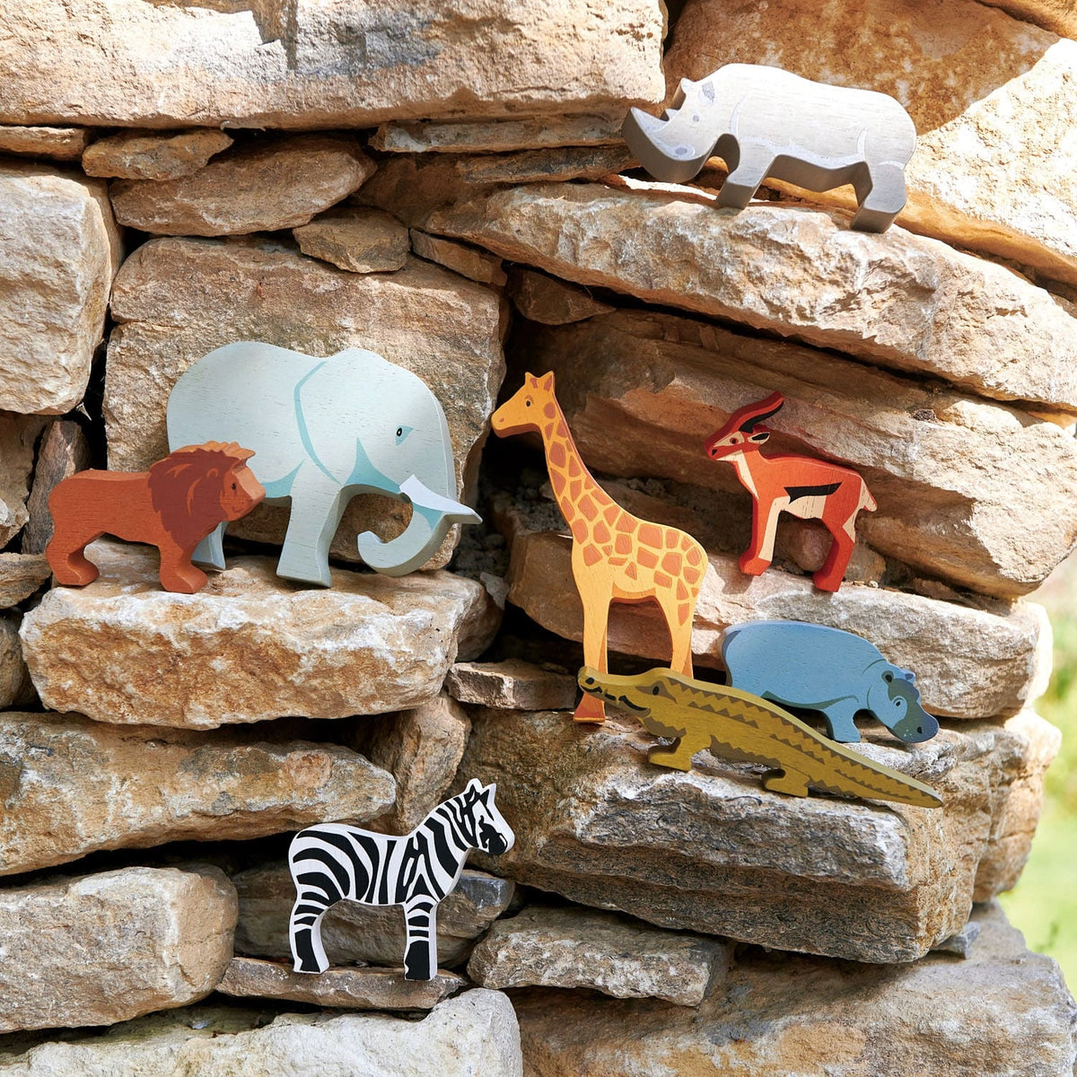tender leaf toys wooden safari animals play figures including alligator, zebra, giraffe, antelope, lion, elephant, rhino and hippo on a stone wall outside