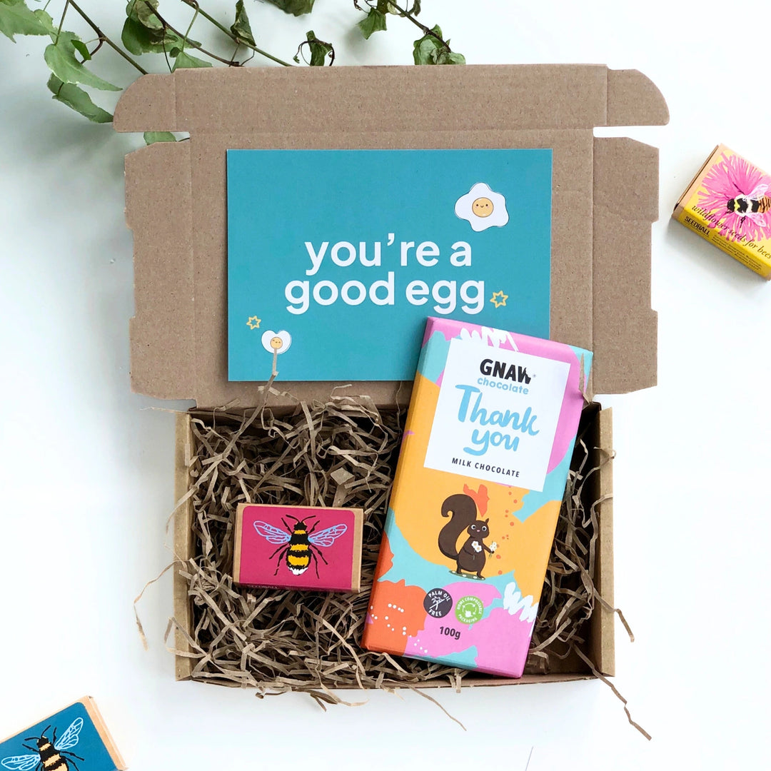 kraft gift box filled with shredded paper and including a coloufyl thnak you chocolate bar and a box of bee friendly seedballs. The gift box comes with a teal green card with the phrase 'you're a good egg' in bold white text. 