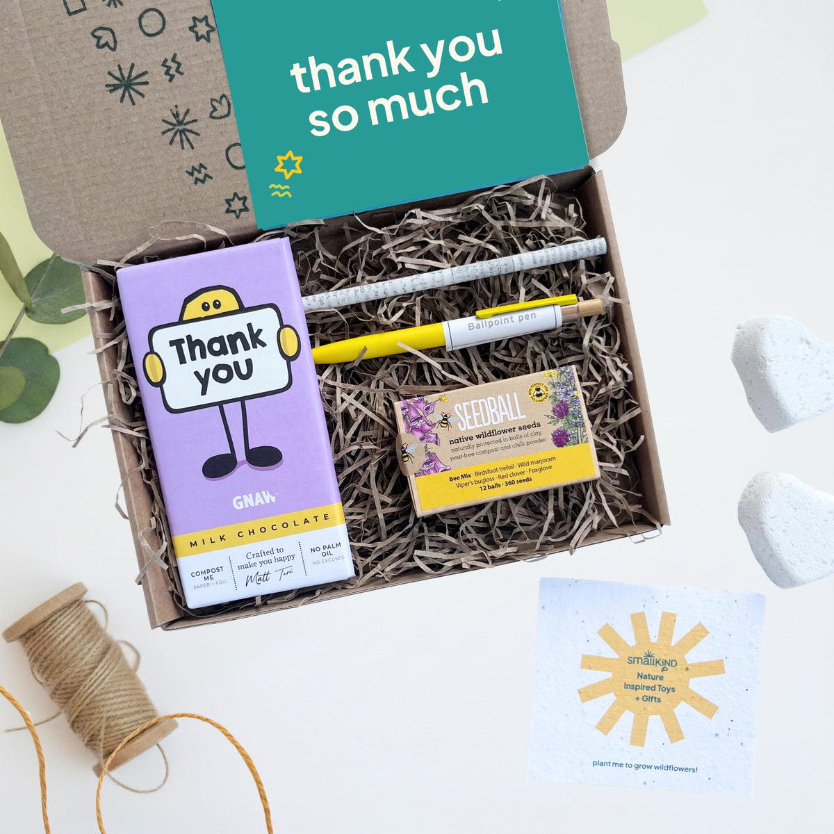 Gift box with chocolate, pen, pencil and seed box labeled 'thank you so much' on a light background.
