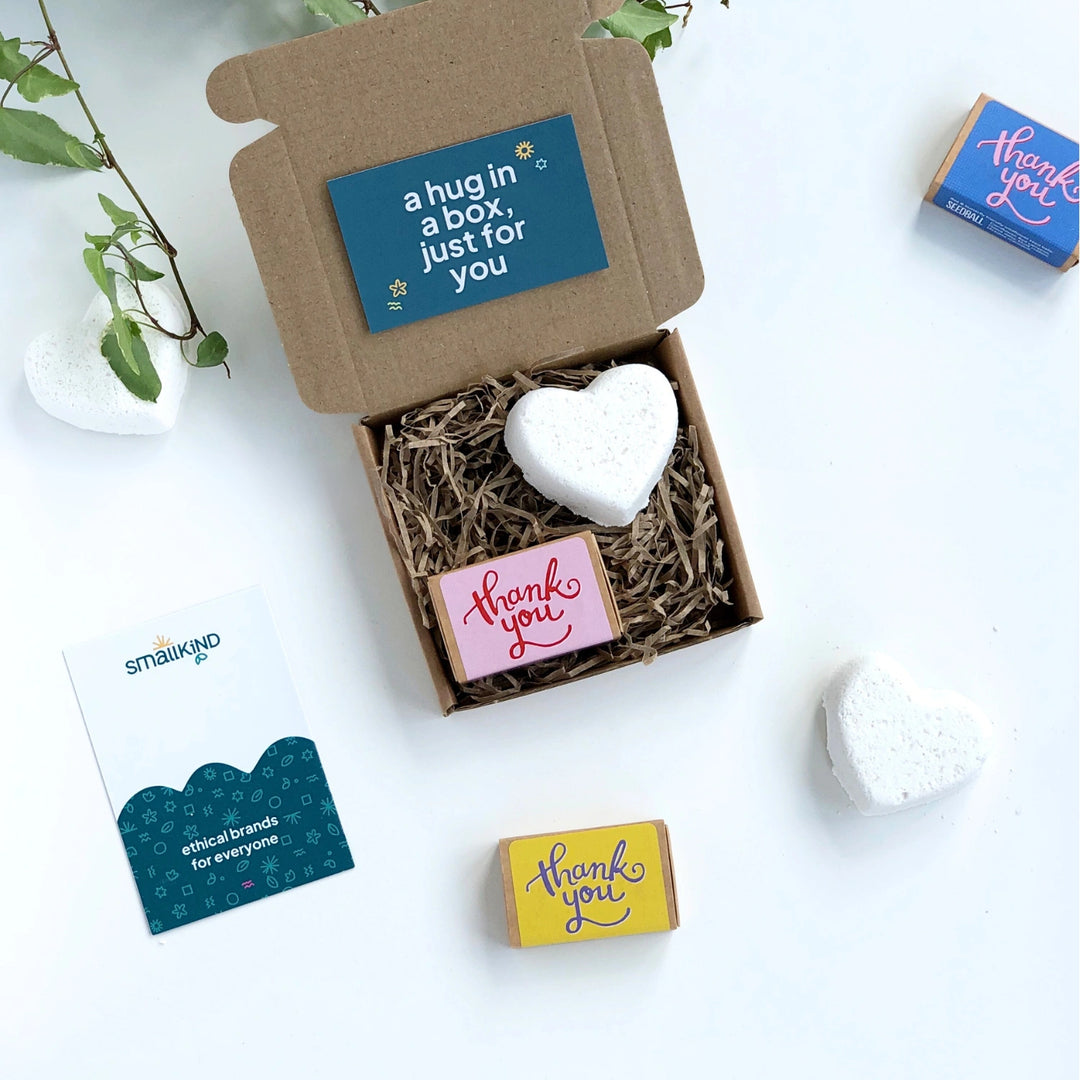 a white heart shaped bath bomb and matchbox sized box of seedballs inside a small kraft gift box filled with shredded paper. 