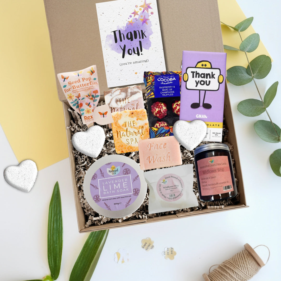 Gift box with various self care products including a face wash, soap, and a candle with a thank you card on a white background.