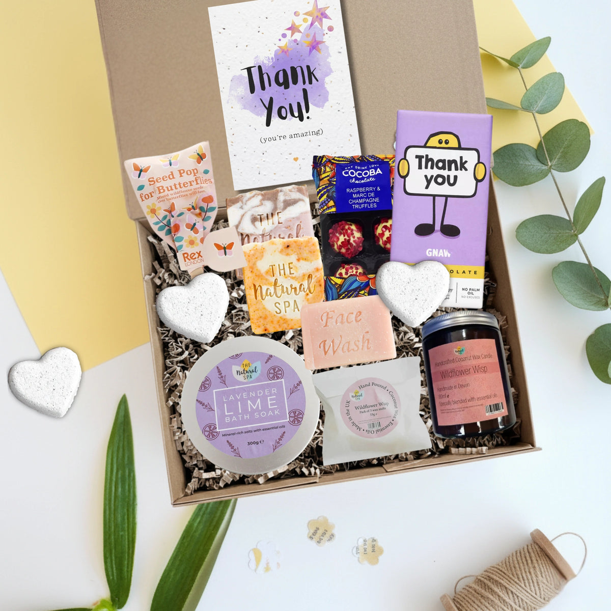 Gift box with various self care products including a face wash, soap, and a candle with a thank you card on a white background.