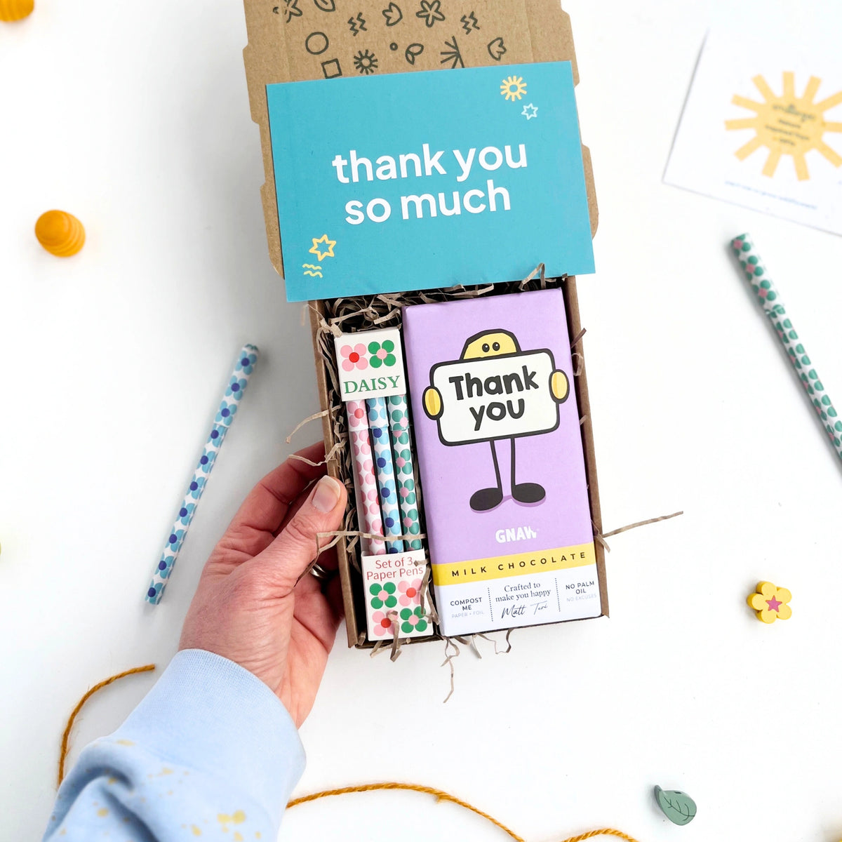 hand holding a smallkind thank you treat box with thank you chocolate bar in purple wrapper with a set of three eco friendly paper pens in a daisy design. 