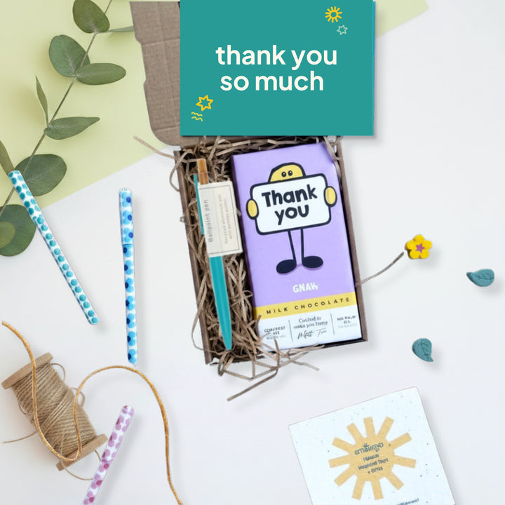 A gift box containing a purple 'Thank You' milk chocolate bar, styled on a white surface with decorative props including a green leafy branch, patterned pens, twine, and a small card. A teal card at the top reads 'thank you so much'.