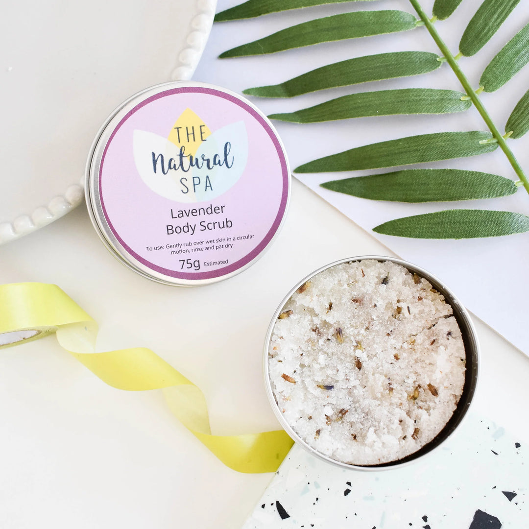 An open round tin of body scrub next to its labeled lid on a white surface, styled with green leaves and a yellow ribbon. The label reads &