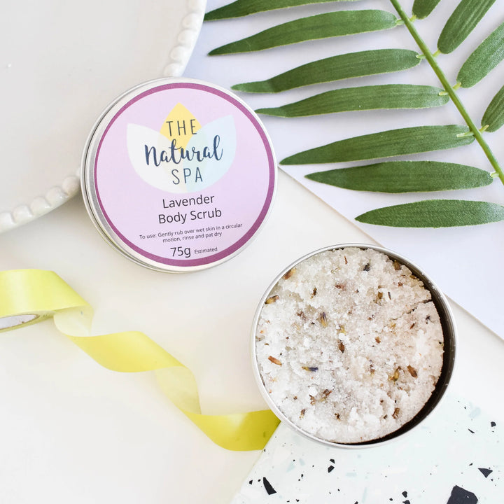 An open round tin of body scrub next to its labeled lid on a white surface, styled with green leaves and a yellow ribbon. The label reads &