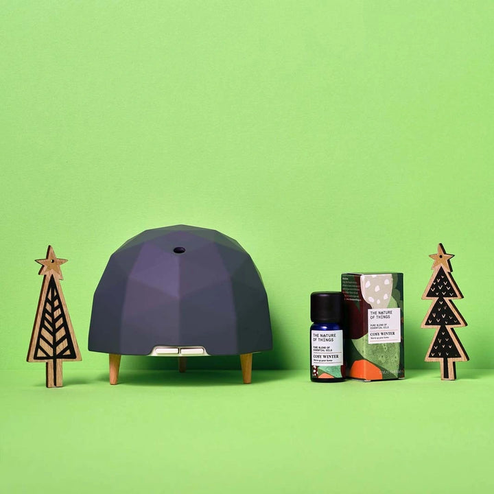 Cosy Winter Essential Oil Blend with a grey dome shaped diffuser and christmas tree decorations on a green background 