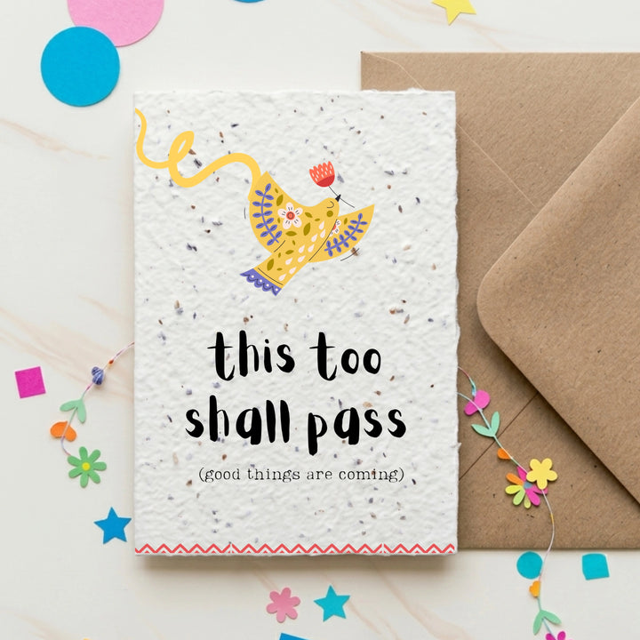 Motivational card with 'this too shall pass' text and a yellow bird illustration, placed on a white surface with colourful confetti.