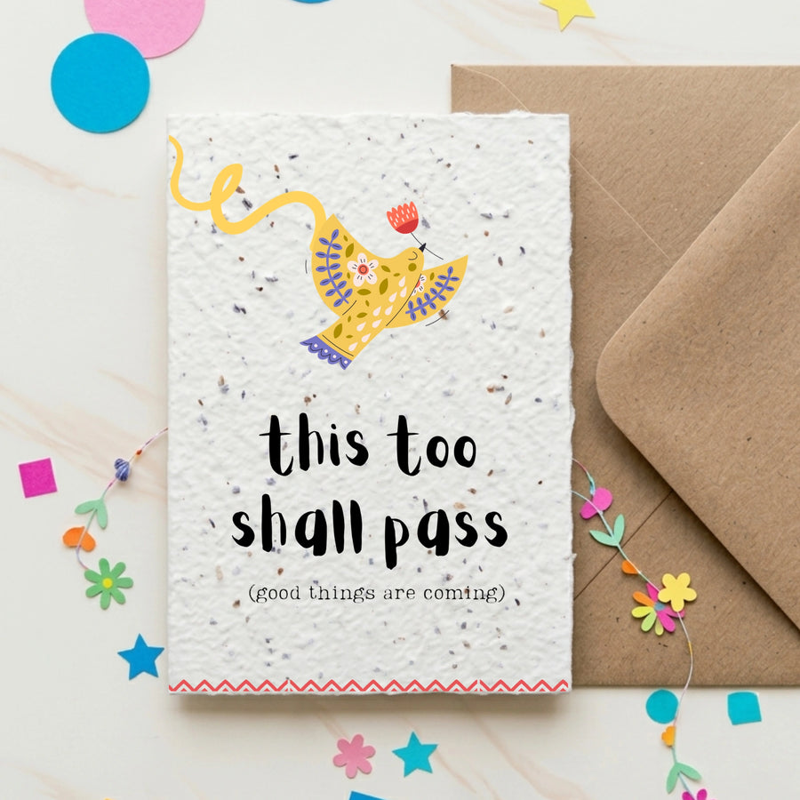 Motivational card with 'this too shall pass' text and a yellow bird illustration, placed on a white surface with colourful confetti.