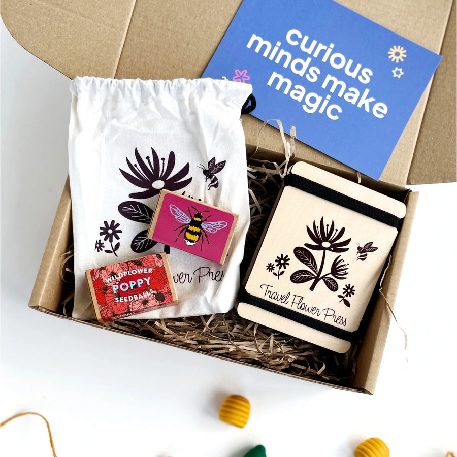 travel size wooden flower press with a cotton storage bagg and two boxes of wildflower seeds. The products are inside a kraft gift box filled with shredded paper and including a blue card which reads 'curious minds make magic'. 