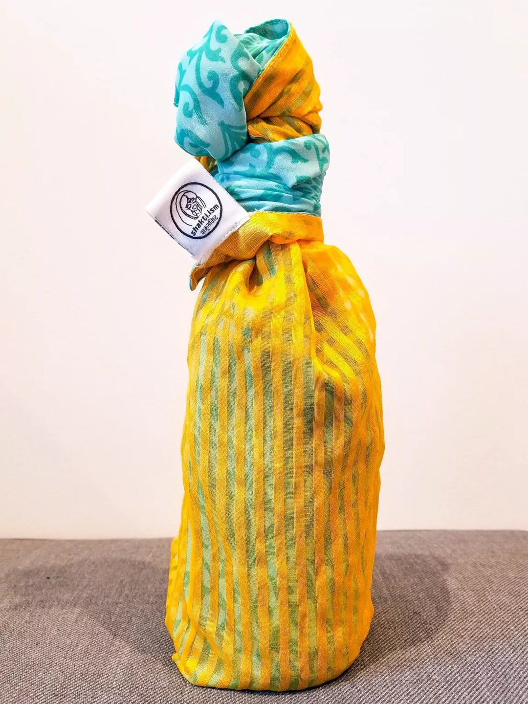 Yellow bottle wrapped in patterned fabric gift wrap with a white label on a grey surface.