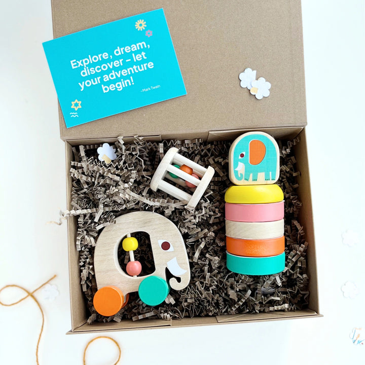 Box of children's toys with a colourful elephant-shaped push along toy, wooden stacker and rattle in a gift box on a white background