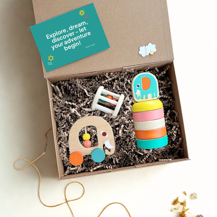 Box of children's wooden toys with a colourful elephant rattle, matching wooden stacker and a push along elephant toy, featuring a green gift card with the words 'explore, dream, discover'.