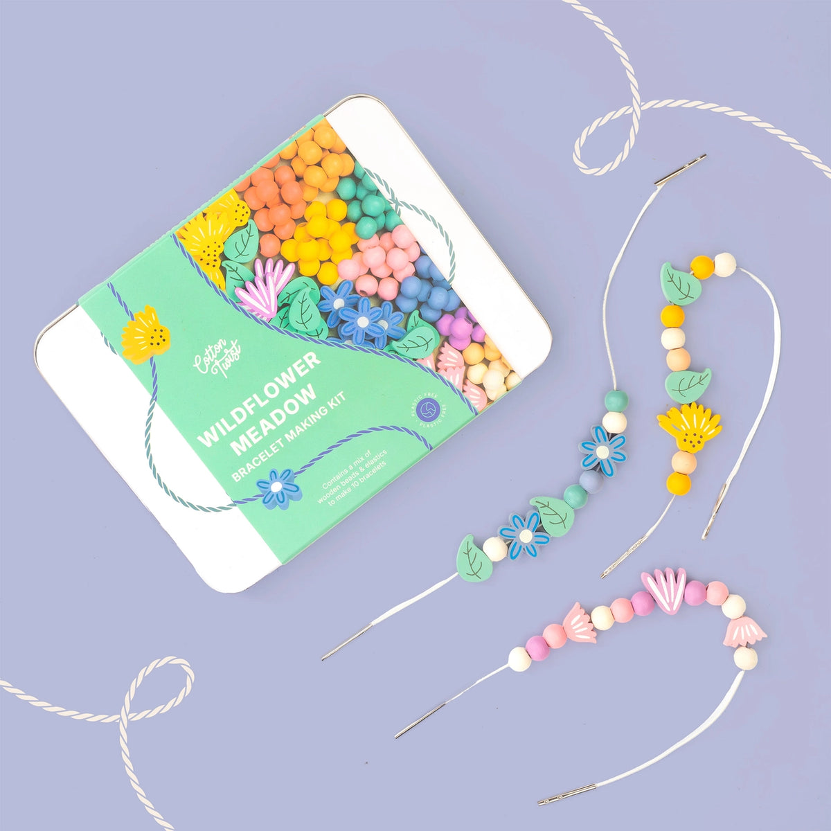 A wildflower meadow bead and stringing kit shown with its mint-green and white tin packaging on a lavender background, alongside several completed stringed bead arrangements made from colorful flower-shaped beads and white cord.