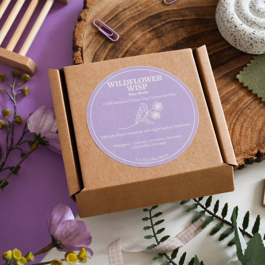 Box of Wildflower Wisp wax melts with a purple label on a wooden surface with flowers and ribbons.
