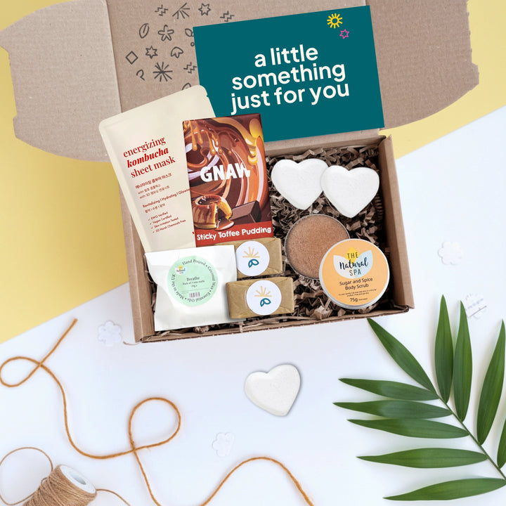 winter self care box with various items including a face mask, soaps, chocolate and heart-shaped bath bombs on a light background