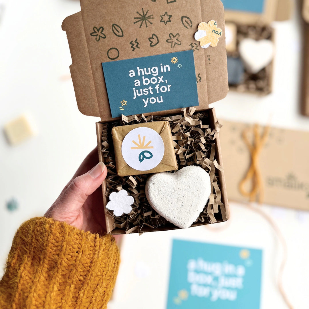 womans hand holding a Hug in a box with Heart-shaped bath bomb and wrapped mini soap bar in a gift box with seed paper shapes, shredded paper and a 'hig in a box' gift card. 