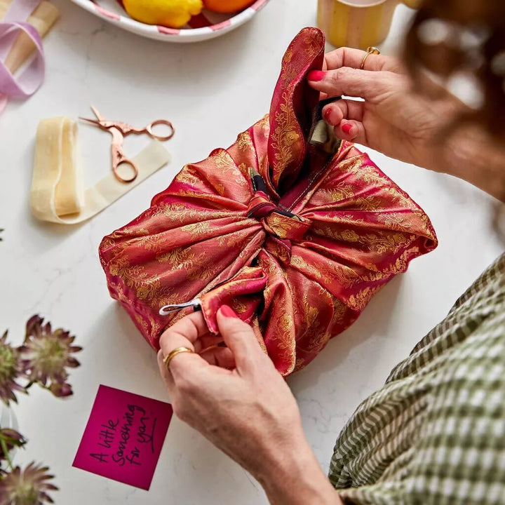 Person wrapping a gift in red and gold fabric with scissors and flowers on a table.