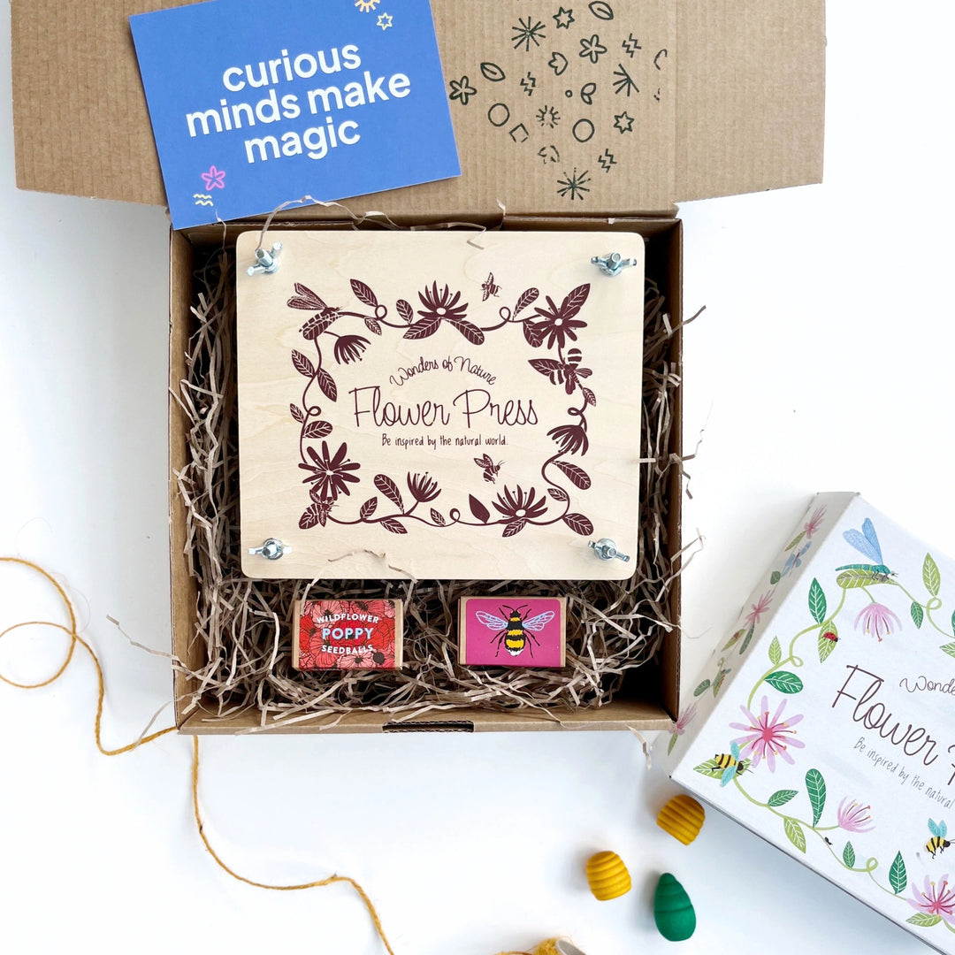 wooden flower press with floral engraving and two boxes of wildflower seedballs in a hand stamped kraft gift boxed filled with shredded paper. The gift boxes comes with a card that reads 'curious minds make magic'. 