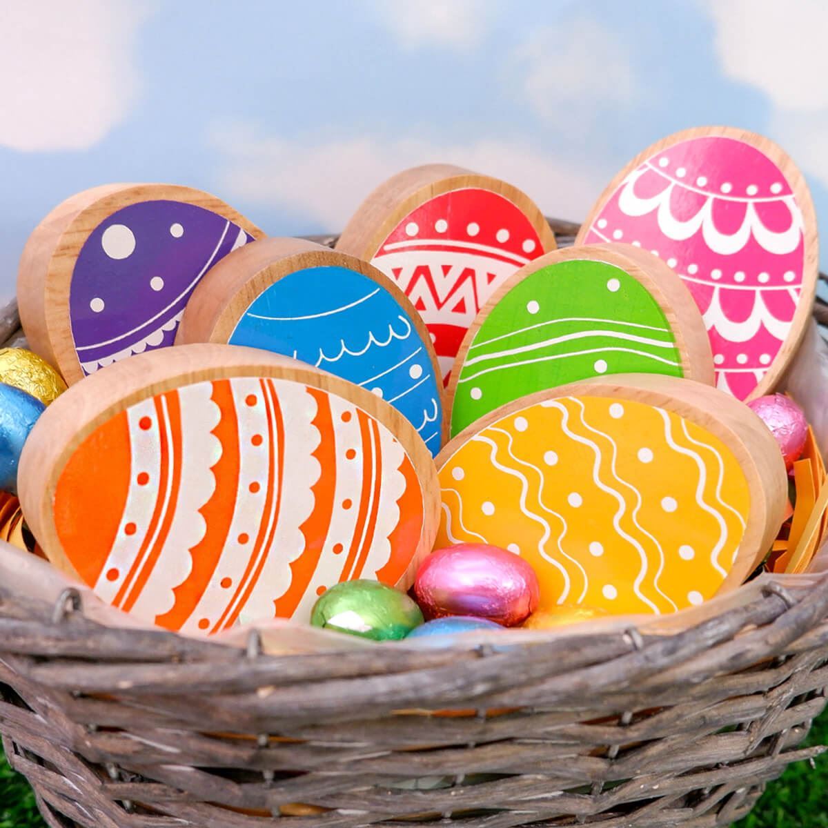 wooden easter eggs play figures with colourful painted details and a natural wood grain edge. The eggs are in a wicker basket with chocolate easter eggs.