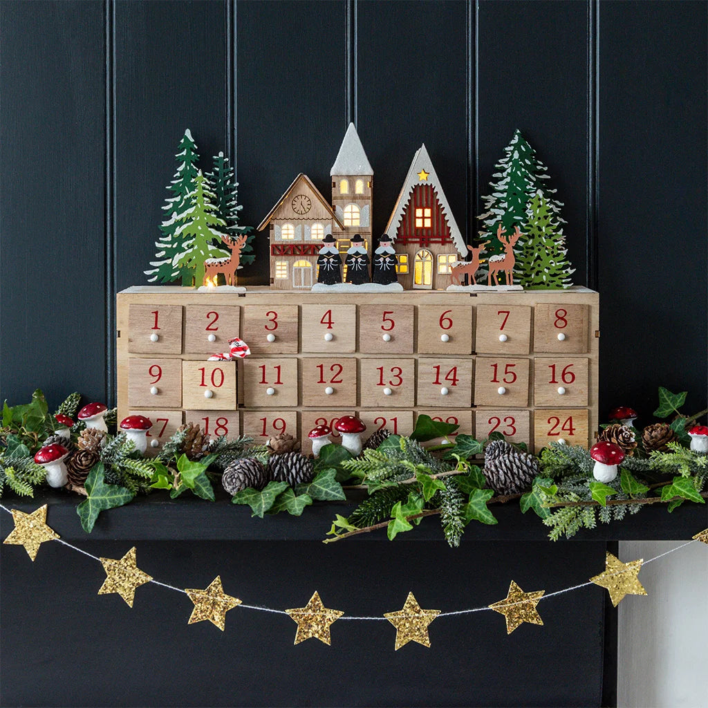 A wooden Christmas advent calendar with 24 numbered drawers, decorated with miniature winter houses, trees, deer, pinecones, red-and-white mushrooms, and green foliage garlands, set against a dark paneled background.