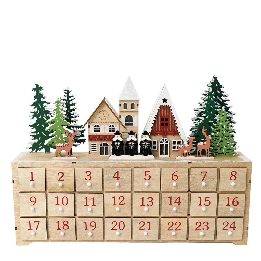 A wooden Advent calendar with 24 numbered drawers on the front, decorated with small Christmas houses, evergreen trees, and deer on top, photographed against a plain white background.