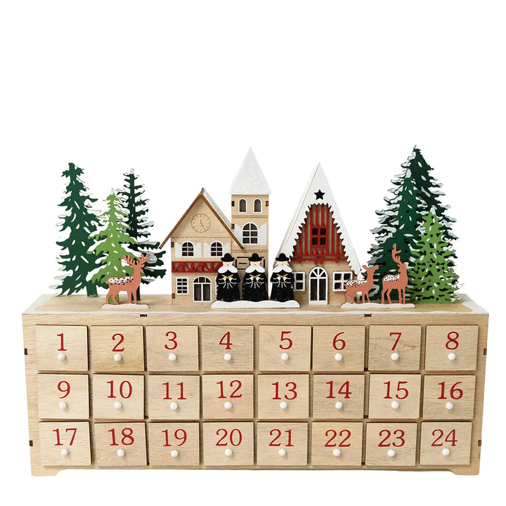 A wooden Advent calendar with 24 numbered drawers on the front, decorated with small Christmas houses, evergreen trees, and deer on top, photographed against a plain white background.