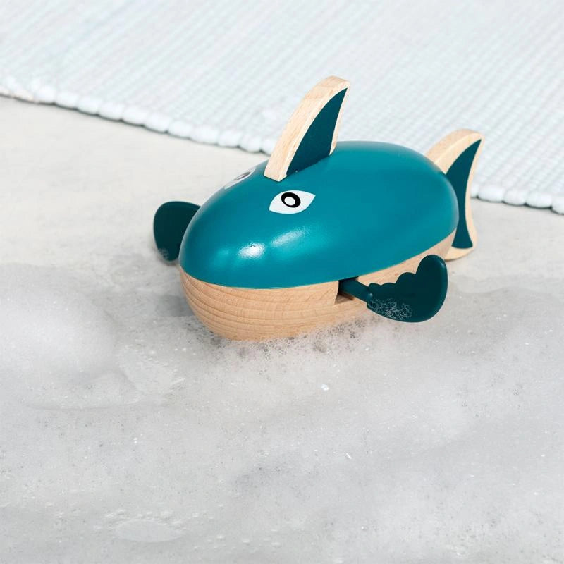 teal green wooden wind up shark bath toy on a light coloured bathroom surface with bubbles and bath foam